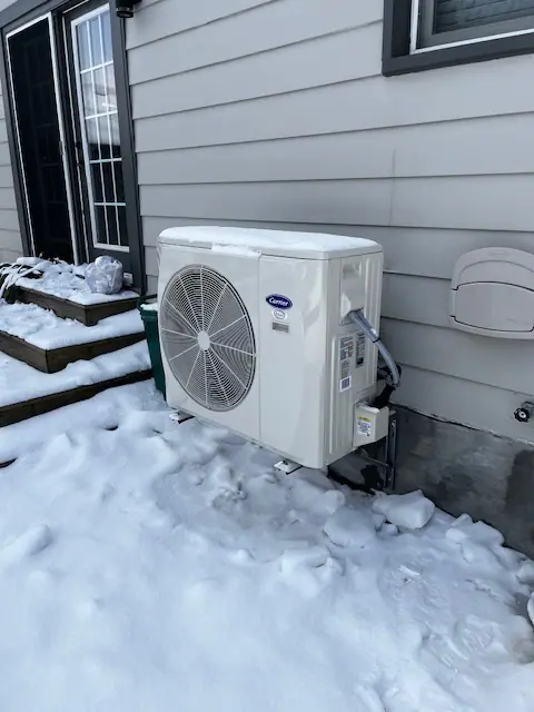 Carrier outdoor heat pump on a raised base beside a house in snowy conditions